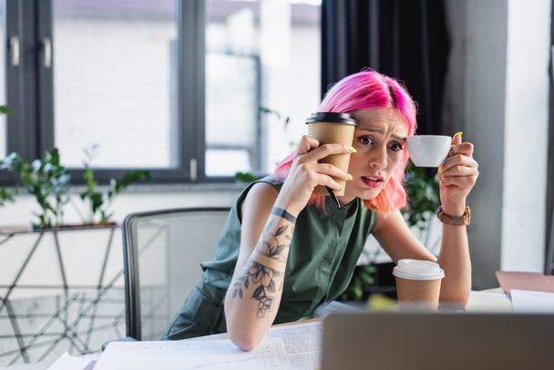στρεσαρισμένη επιχειρηματίας με ροζ μαλλιά κρατώντας καφέ και κοιτάζοντας το laptop  - Φωτογραφία, εικόνα