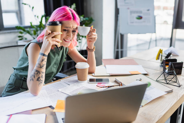 worried businesswoman with pink hair holding coffee and looking at laptop  - Fotó, kép