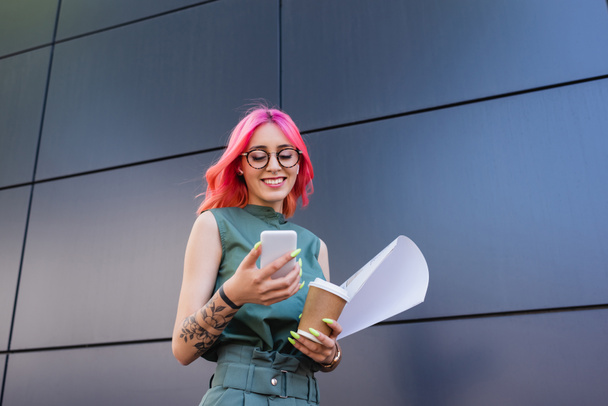 happy and tattooed businesswoman in earphones using smartphone while holding paper cup - Photo, Image