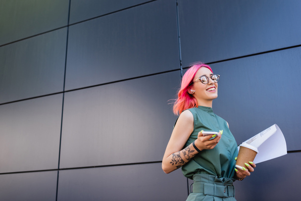 happy and tattooed businesswoman in earphone holding smartphone and paper cup - Fotoğraf, Görsel