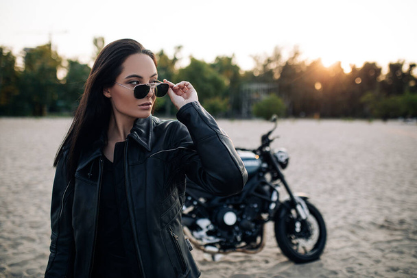 Young woman stands in sunglasses and leather jacket among sandy beach against background of black motorbike at sunset. - Foto, imagen