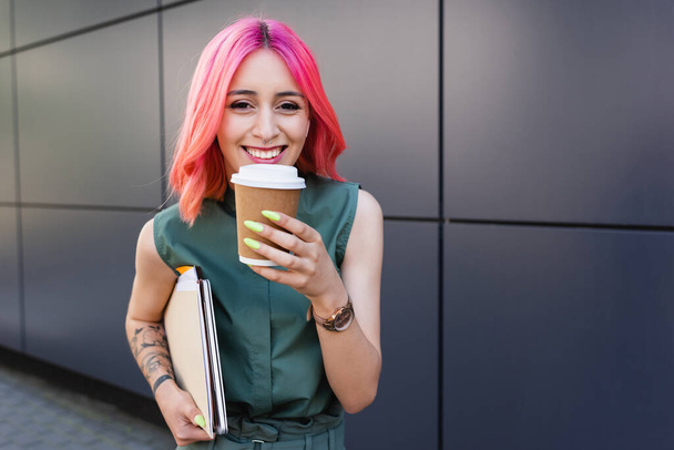 happy businesswoman with pink hair and earphone holding coffee to go and folders - Foto, immagini