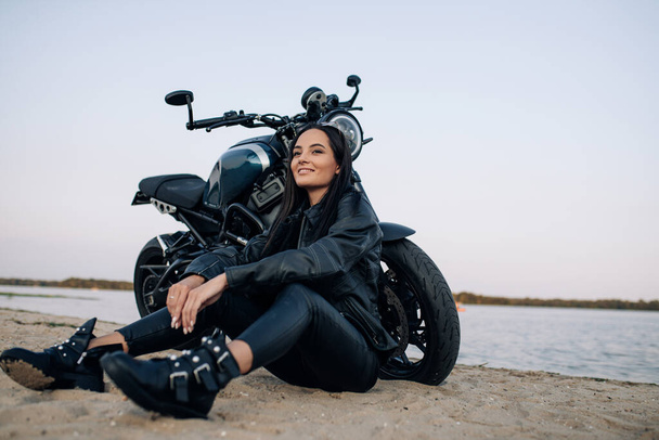 Młoda kobieta siedzi na piasku w pobliżu czarnego motocykla w skórzanej kurtce i spodniach na plaży nad brzegiem rzeki. - Zdjęcie, obraz