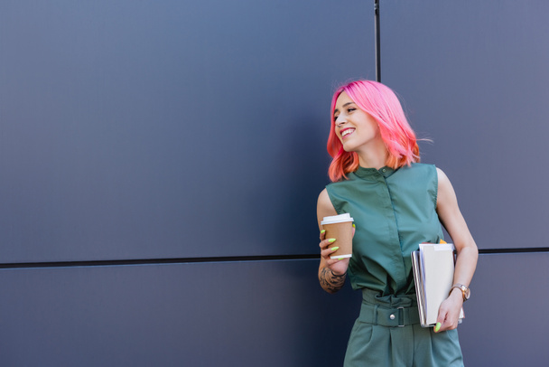 tattooed and smiling businesswoman with pink hair holding folder and coffee to go outside - Fotó, kép