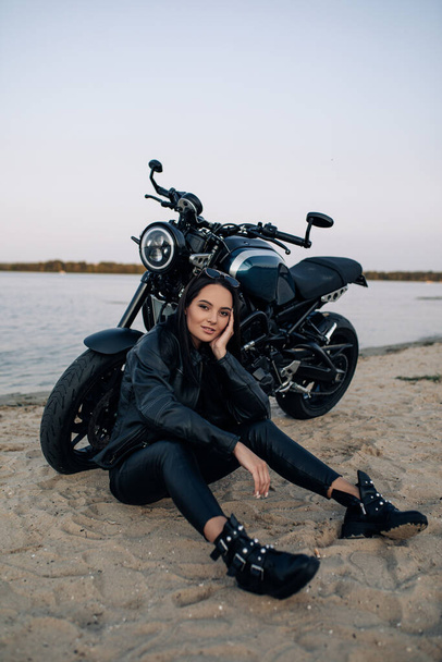 Young woman sits on sand near black motorbike in leather jacket and pants on beach near river coast. - Фото, изображение