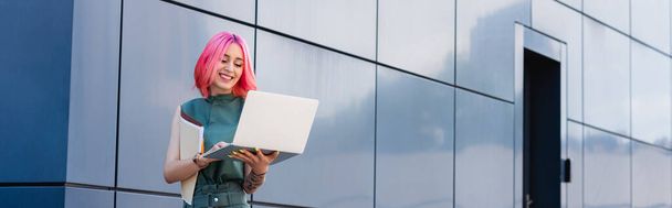 femme d'affaires tatouée et positive avec des cheveux roses tenant ordinateur portable et dossier à l'extérieur, bannière - Photo, image