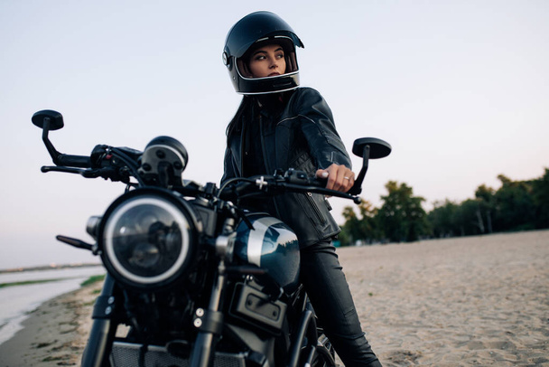 Young woman sits on black motorbike in helmet and leather jacket on sandy beach next coast of river. - Foto, afbeelding