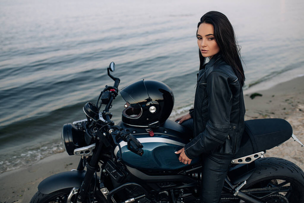 Молода жінка сидить на чорному мотоциклі в шкіряній куртці і штанях на піщаному пляжі біля узбережжя річки
. - Фото, зображення