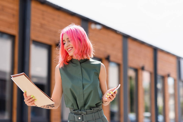donna d'affari soddisfatta con cartelle rosa per capelli e smartphone all'esterno  - Foto, immagini