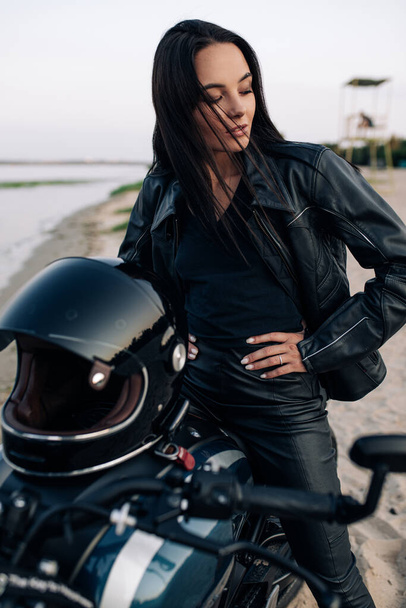 Young woman sits on black motorbike in leather jacket and pants on sandy beach next coast of river. Closeup. - Foto, afbeelding