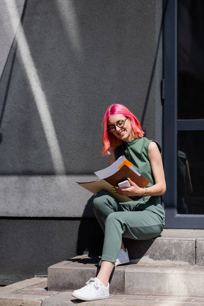 femme d'affaires heureuse avec des cheveux roses tenant dossier avec des documents et smartphone tout en étant assis sur les escaliers à l'extérieur  - Photo, image