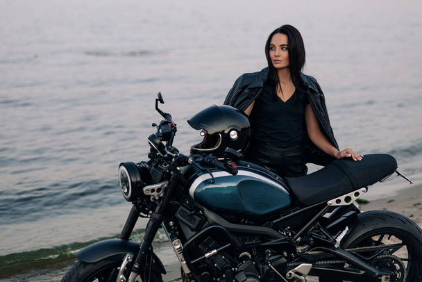 Молода жінка стоїть біля чорного мотоцикла в шкіряній куртці і штанях на піщаному пляжі біля узбережжя річки
. - Фото, зображення
