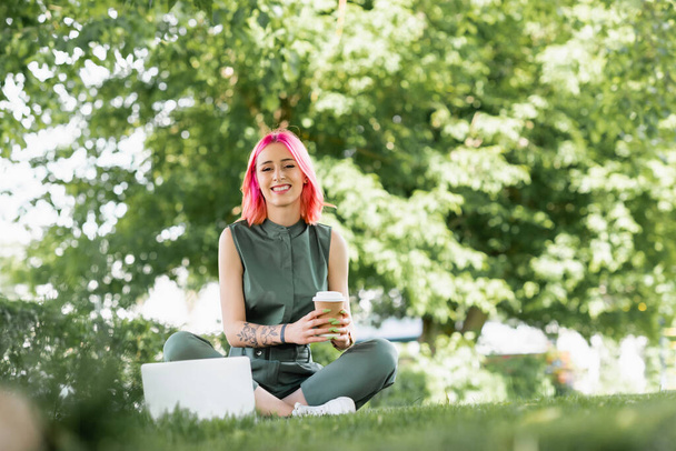 happy young woman with pink hair holding paper cup near laptop on grass  - Foto, Imagem