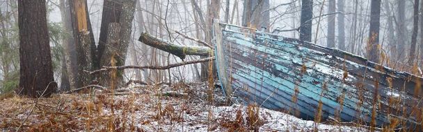 Εγκαταλελειμμένο παλιό μπλε ξύλινο σκάφος σε ένα μυστηριώδες χειμερινό δάσος. Κουνιστά δέντρα σε άσπρη ομίχλη. Φρέσκο χιόνι και χρυσά φθινοπωρινά φύλλα. Concept art, οικονομική παρακμή, ύφεση, φιλοσοφία, αντιθέσεις, παράδοξο - Φωτογραφία, εικόνα