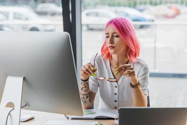 businesswoman with pink hair holding eyeglasses near computer monitor in office - Photo, Image