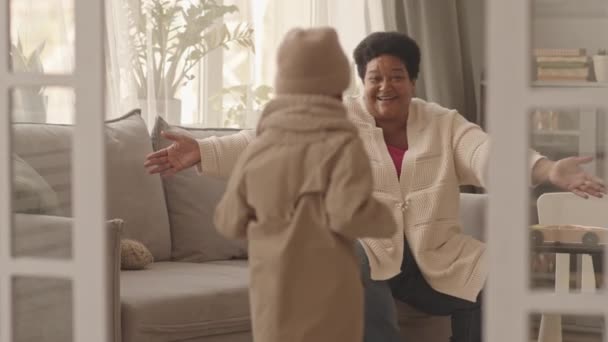Encerrado medio largo de cuidar a la mujer afroamericana mayor sentada en el sofá en la sala de estar durante el día, conociendo y abrazando a su nieto lindo con sombrero de punto y bufanda que llega a casa después de caminar - Imágenes, Vídeo