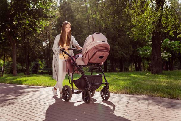 Фотографія повного розміру тіла молода мати посміхається, йдучи в міський парк з дитячою коляскою щасливий
 - Фото, зображення