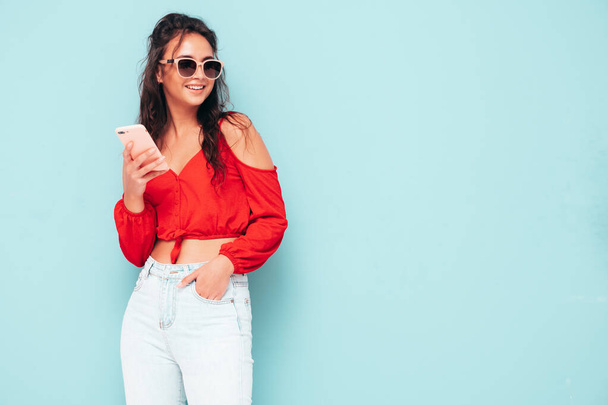 Jovem bela fêmea sorridente na moda verão top vermelho e jeans roupas. Mulher despreocupada Sexy posando perto da parede azul no estúdio. Olhando para a tela do smartphone. Modelo usando aplicativos de celular - Foto, Imagem