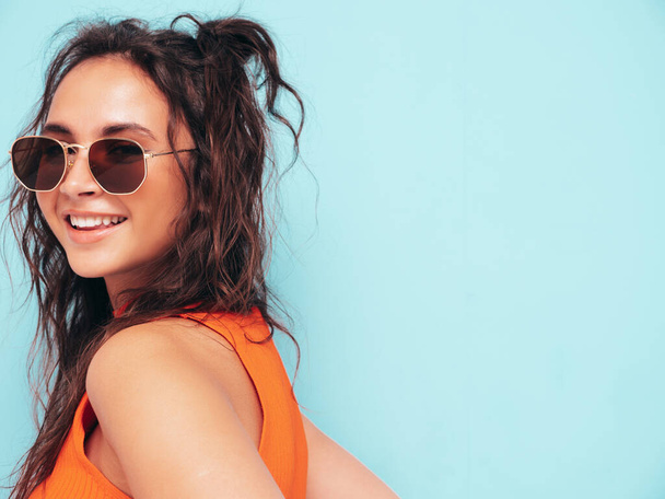 Closeup portrait of young beautiful smiling female in trendy clothes.Sexy carefree woman posing near blue wall in studio.Positive model having fun indoors.Cheerful and happy. In sunglasses - Valokuva, kuva