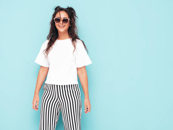 Young beautiful smiling female in trendy summer clothes. Sexy carefree woman posing near blue wall in studio. Positive brunette model having fun and going crazy. Cheerful and happy in sunglasses - Φωτογραφία, εικόνα