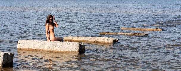 Молодая обнаженная женщина в солнечный летний день загорает на бетонных плитах, лежащих в море
 - Фото, изображение