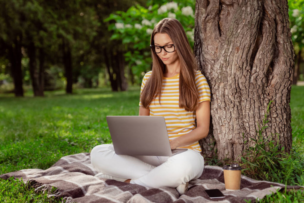 Full length body size photo young woman working on computer in city park wearing glasses sitting on blanket - 写真・画像