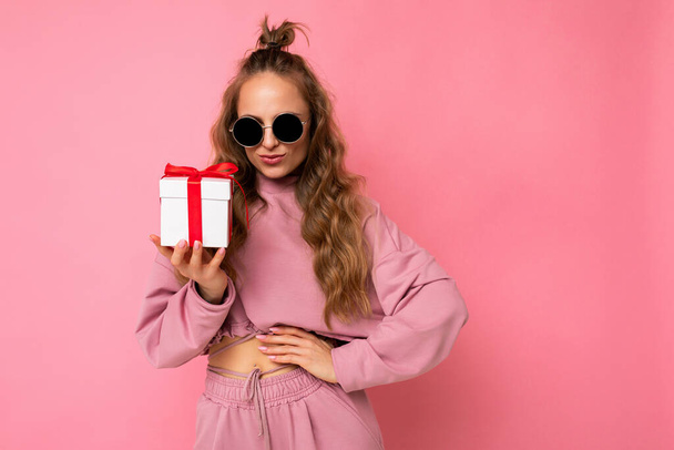 Attractive happy sexy young blonde curly woman isolated over pink background wall wearing pink sport clothes and sunglasses holding gift box looking at camera - Photo, Image