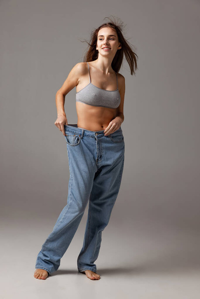 Portrait of young beautiful slim girl in blue jeans jumpersuit posing isolated over gray studio background. Weight Loss concept. - Foto, Imagen