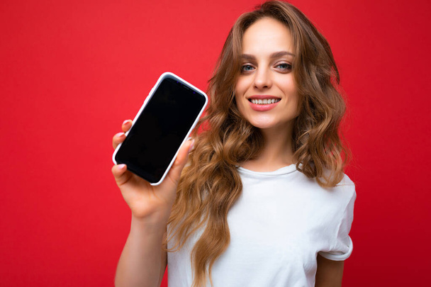 Φωτογραφία της όμορφης χαμογελαστή νεαρή γυναίκα όμορφη φορώντας casual κομψό ντύσιμο στέκεται απομονωμένη στο παρασκήνιο με αντίγραφο χώρο κρατώντας smartphone δείχνει τηλέφωνο στο χέρι με άδεια οθόνη οθόνη για - Φωτογραφία, εικόνα