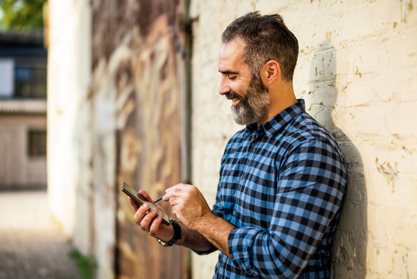 Businessman with a beard using mobile phone with digitized pen while standing in front of wall. - Фото, изображение