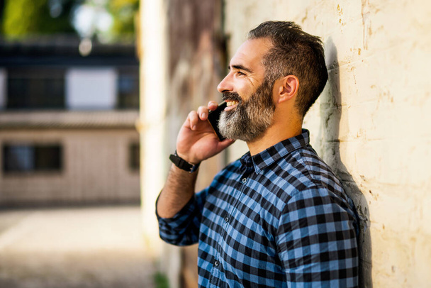 Modern businessman with beard using mobile phone while standing in front of wall outdoor. - Foto, Imagen