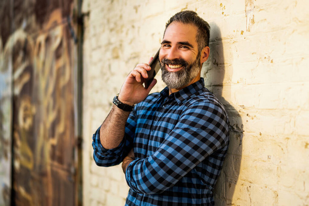 Modern businessman with beard using mobile phone while standing in front of wall outdoor. - Foto, Imagen