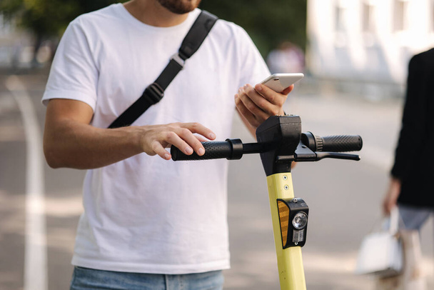 Giovane uomo collegare uno scooter elettrico all'aperto. Uomo con borsa a tracolla nera utilizzando il telefono per il pagamento senza contatto. Concetto di trasporto ecologico - Foto, immagini