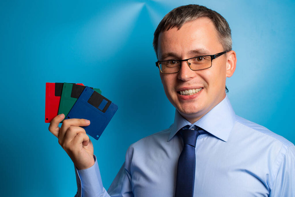 A man in a blue shirt, tie and glasses shows three floppy disks in blue, green and red. Retro technology concept. - Foto, immagini