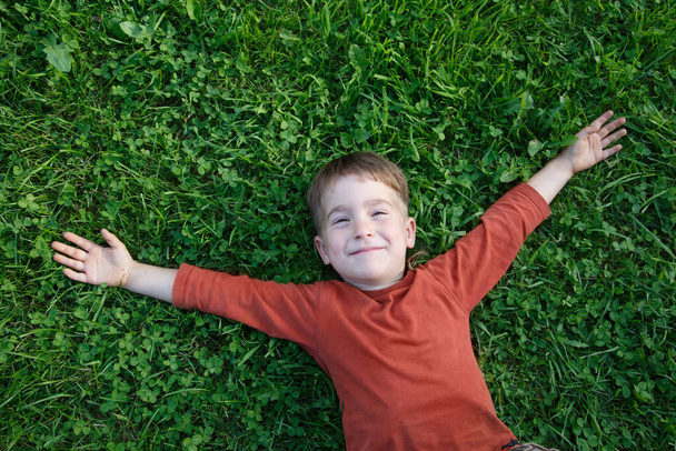 A little cute boy is lying on the green grass with his arms outstretched joyfully. - Foto, Imagen