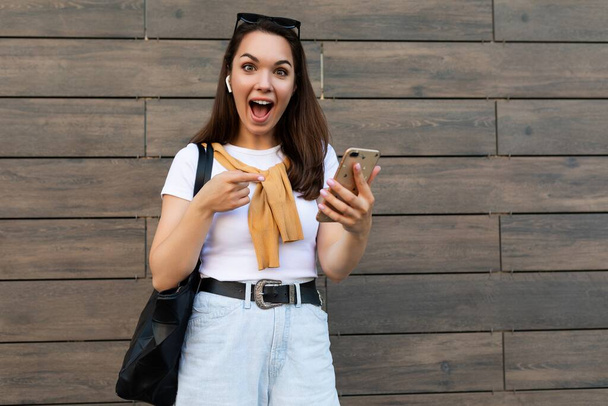Фото привабливої шокованої здивованої молодої жінки в повсякденному одязі, що стоїть на вулиці, використовуючи мобільний телефон, дивлячись на камеру
 - Фото, зображення