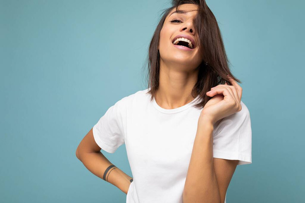Photo of young beautiful happy smiling brunette woman wearing trendy white t-shirt with empty space for mock up. Sexy carefree female person posing isolated near blue wall in studio with free space - 写真・画像
