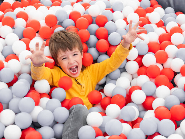 Laughing boy plays in ball pit. White and red plastic balls in dry paddling pool. Leisure activity in playroom at kindergarten. Leisure activity. Active recreation. - Foto, Bild