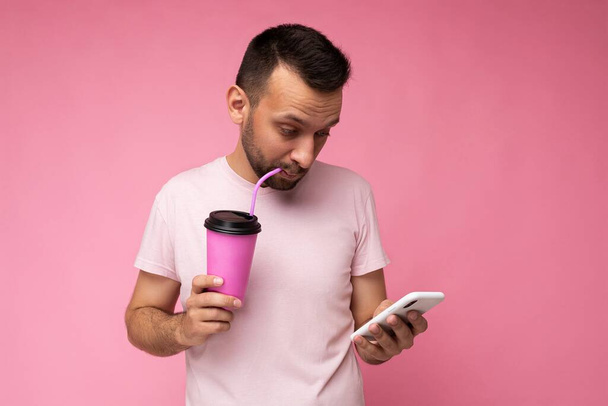 Όμορφος χαρούμενος μελαχροινή αξύριστος άντρας με γενειάδα φορώντας λευκό t-shirt που απομονώνονται πάνω από ροζ backgroung τοίχο χρησιμοποιώντας κινητό τηλέφωνο κρατώντας χάρτινο φλιτζάνι καφέ για mock up και πόσιμο κοιτάζοντας gadjet - Φωτογραφία, εικόνα