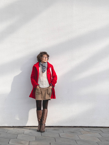 Curly woman in red duffle coat is standing by white wall with sunbeams. Smiling student. Modern lifestyle. - Фото, зображення