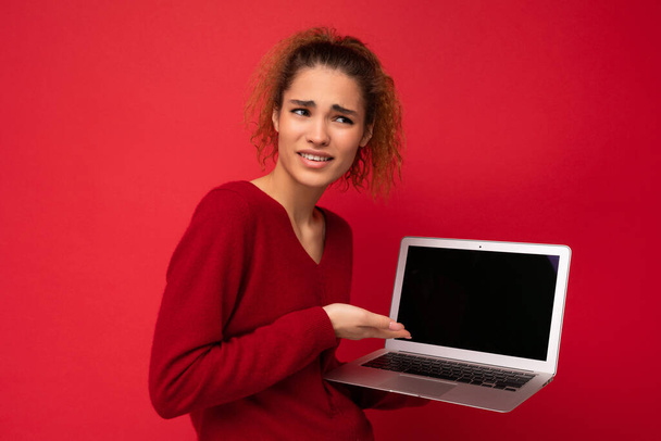 Крупный план портрет красивой удивлённой, удивлённой темноволосой женщины, держащей ноутбук, смотрящий в сторону, показывающий на нетбуке клавиатуру в красном свитере, изолированном над красной стеной - Фото, изображение