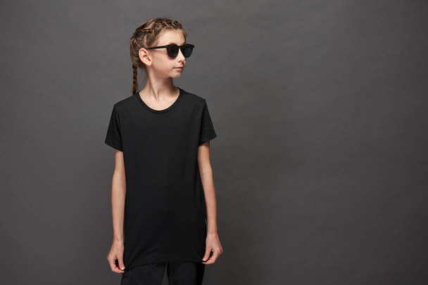 Kid girl wearing black t-shirt with space for your logo or design in studio over gray background with sunglasses - 写真・画像