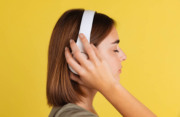 Side view of young cute girl, student in white headphones listening to music isolated on yellow studio backgroud. - Foto, Bild