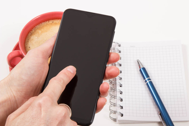 Akıllı telefonlu bir kadın eli, not tutmak için sütlü bir fincan kahve ve not defteri. İş ya da cep telefonuyla rahatlama. Beyaz arkaplan - Fotoğraf, Görsel