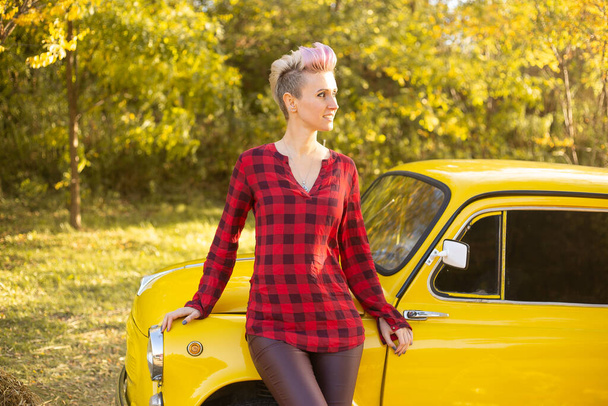 黄金の木と秋の公園の背景に黄色の車の近くに短い髪のヒップスター女性 - 写真・画像