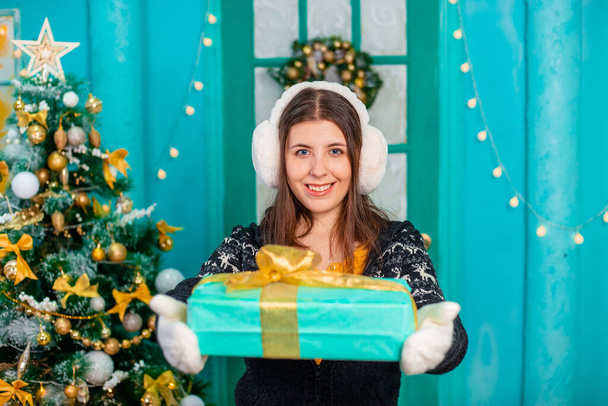 A young, smiling woman dressed in a sweater, warm headphones and mittens is posing with a gift in her hands while standing on the background of the christmas tree. - Fotografie, Obrázek