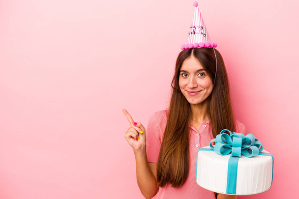 Joven mujer caucásica celebrando un cumpleaños aislada sobre fondo rosa sonriendo y señalando a un lado, mostrando algo en espacio en blanco. - Foto, imagen