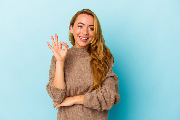 Caucasian woman isolated on blue background winks an eye and holds an okay gesture with hand. - Photo, Image