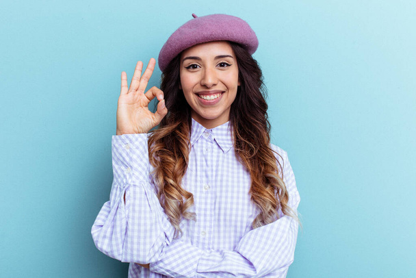 Jeune femme mexicaine isolée sur fond bleu claque un œil et tient un geste correct avec la main. - Photo, image