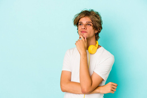 Jonge blanke man luisteren naar muziek geïsoleerd op blauwe achtergrond kijken zijwaarts met twijfelachtige en sceptische uitdrukking. - Foto, afbeelding